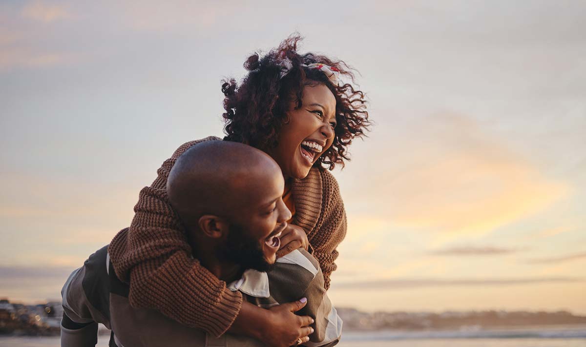 Joyful African-American couple, experiencing the positive outcomes of couples coaching, engaging in playful piggyback riding on the beach, strengthening their bond and communication.